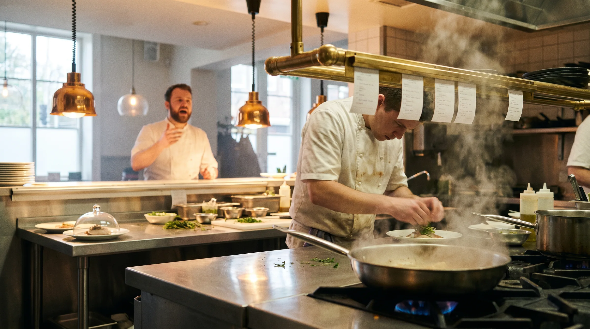 Line cook plating during dinner service in a professional restaurant kitchen, steam rising from a saute pan under brass ticket rail and warm copper pendants.